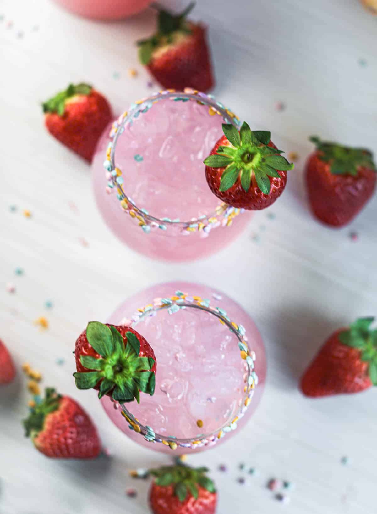 Pink cocktails from above with sprinkles on rim with strawberries and sprinkles on table.