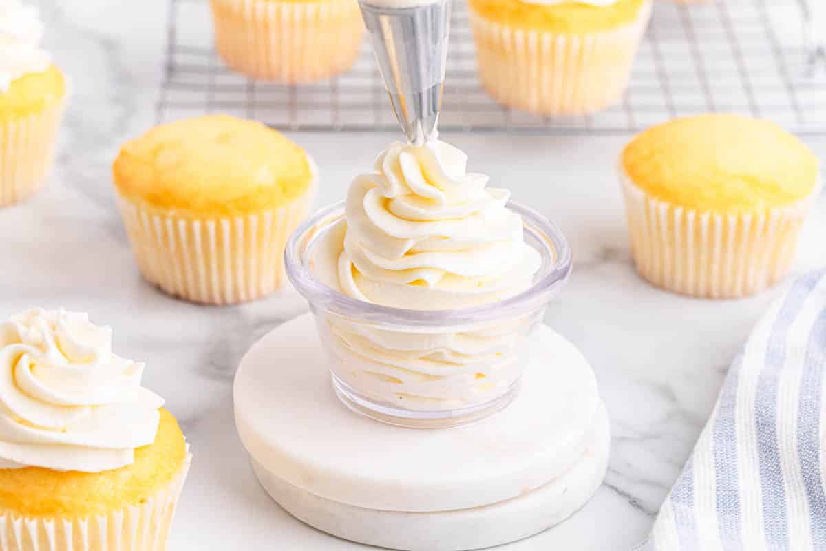 Whipped cream being piped into a glass bowl with cupcakes in background.