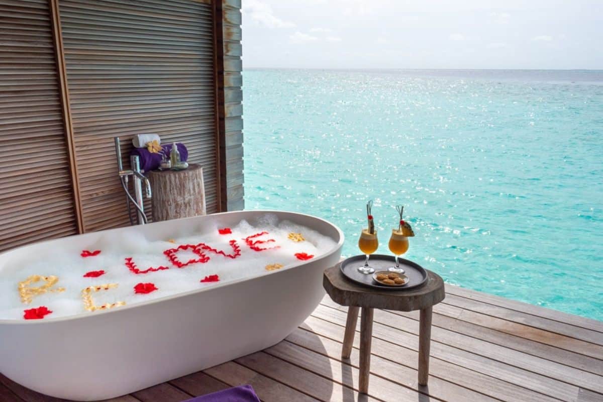 White claw-foot bathtub with Love spelled out in flower petals looking over ocean with cocktails on table.