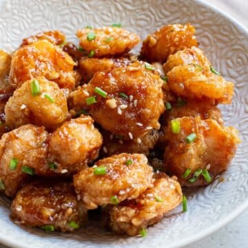 Fried shrimp with sauce and sesame seeds and chives in white bowl with chopsticks.