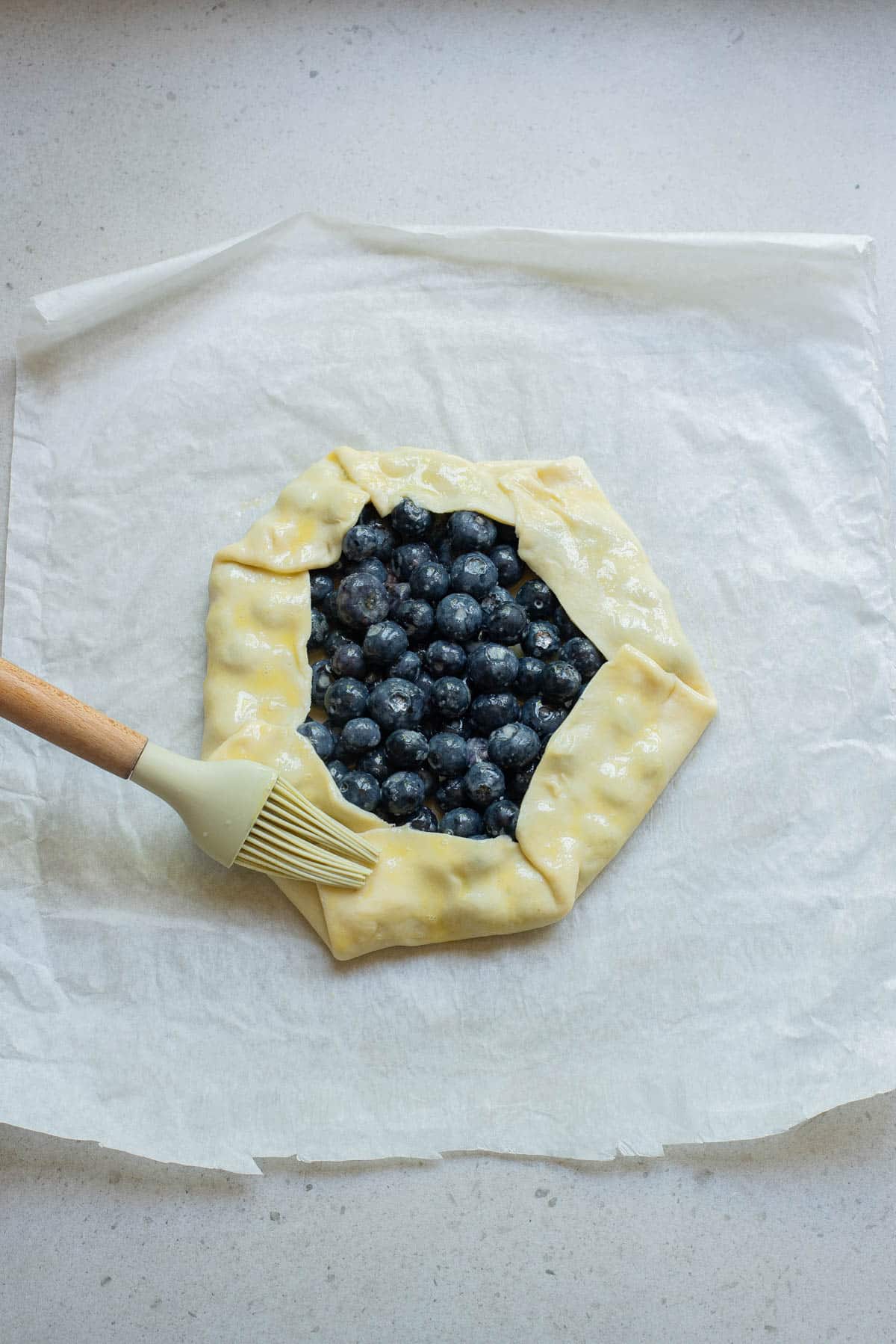 Blueberries in a pie crust on parchment paper brushing with egg wash.