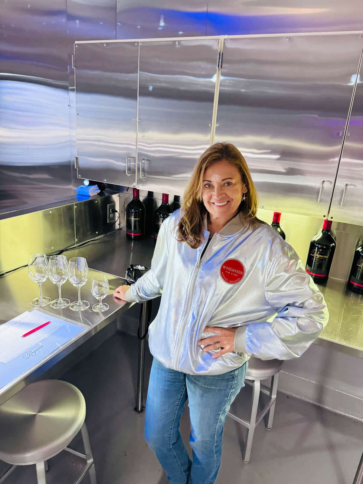 A woman in a silver jacket at a stainless counter with wineglasses and wine in the background.