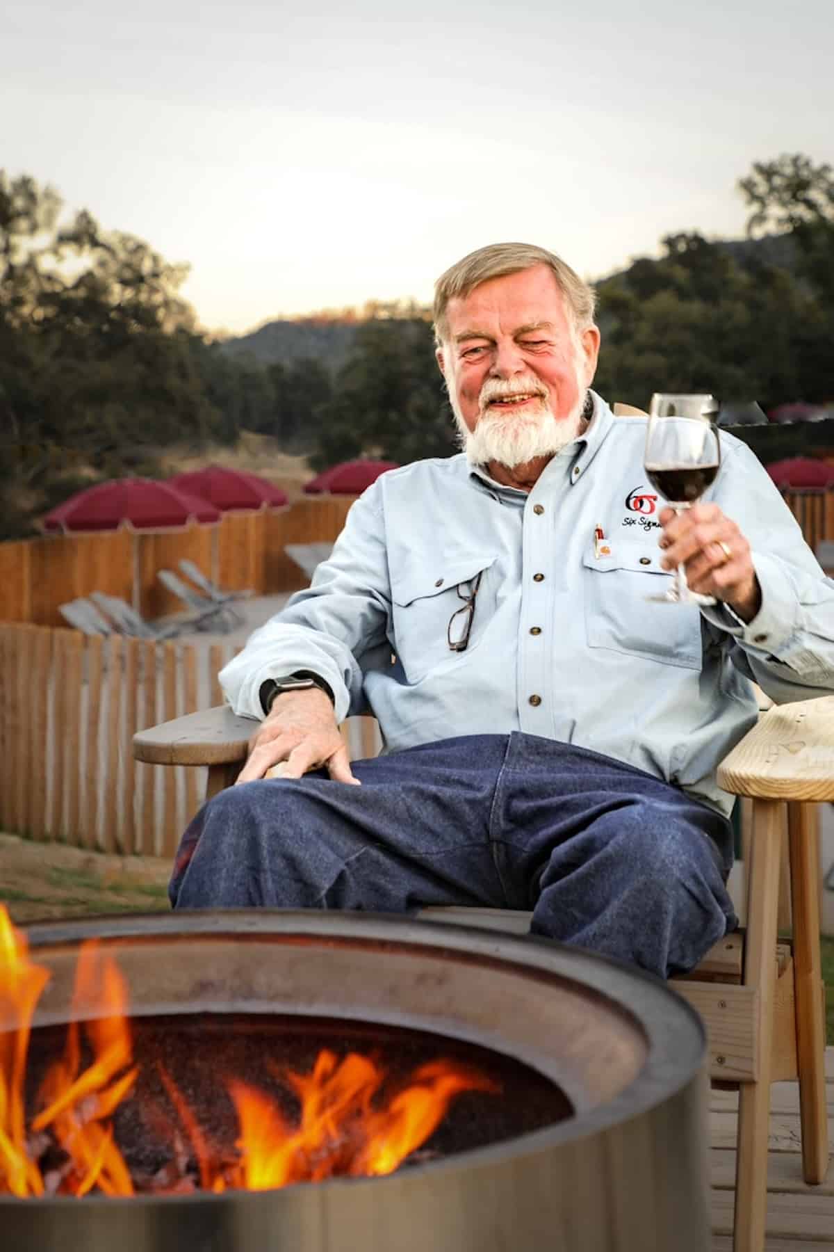 Man seated in front of a fire pit holding a glass of red wine.