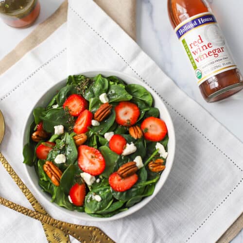 Spinach with strawberries, goat cheese, pecans, and red wine vinaigrette in a white bowl on a white napkin with bottle of red wine vinegar and bronze serving set.