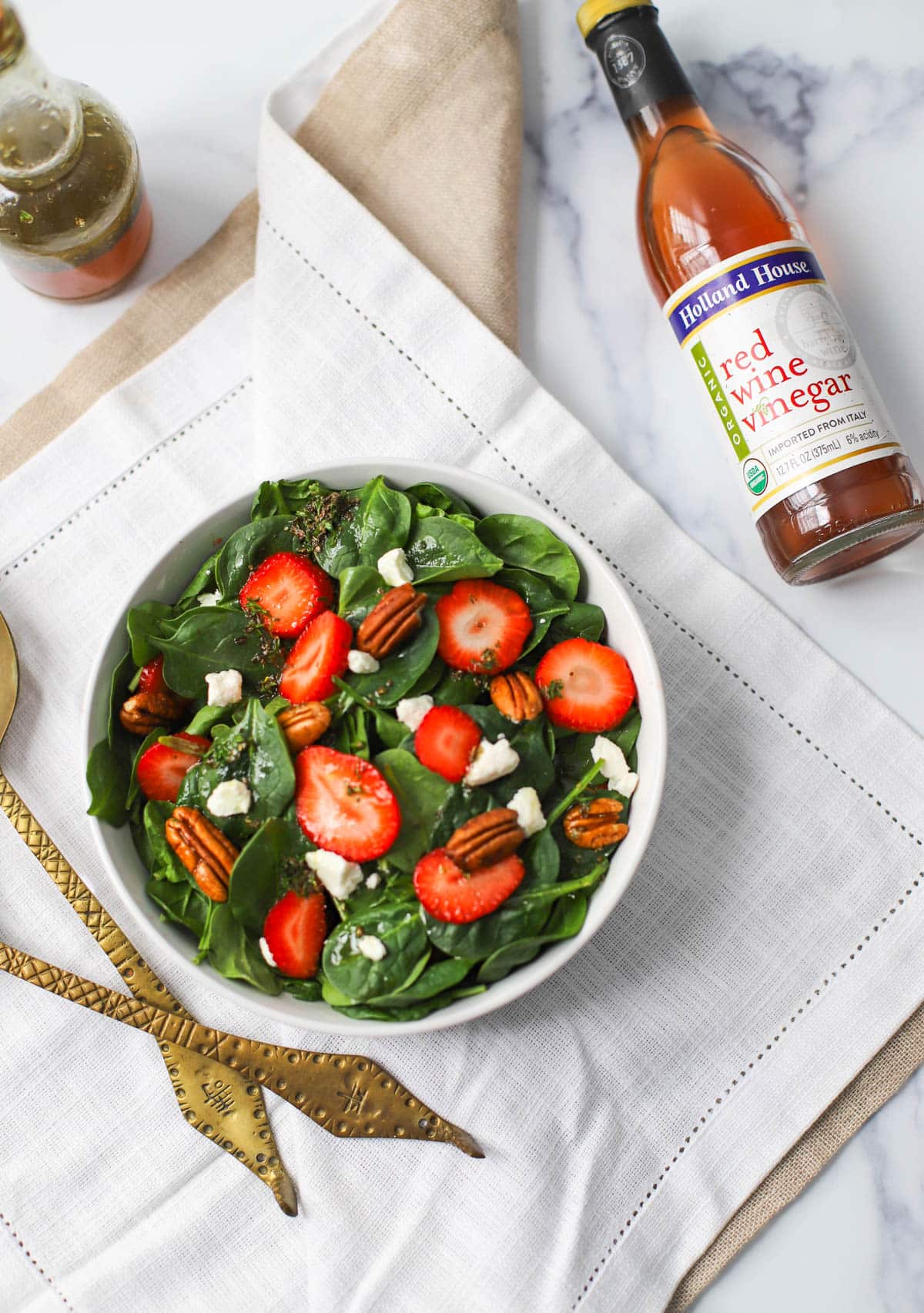 Spinach with strawberries, goat cheese, pecans, and red wine vinaigrette in a white bowl on a white napkin with bottle of red wine vinegar and bronze serving set.