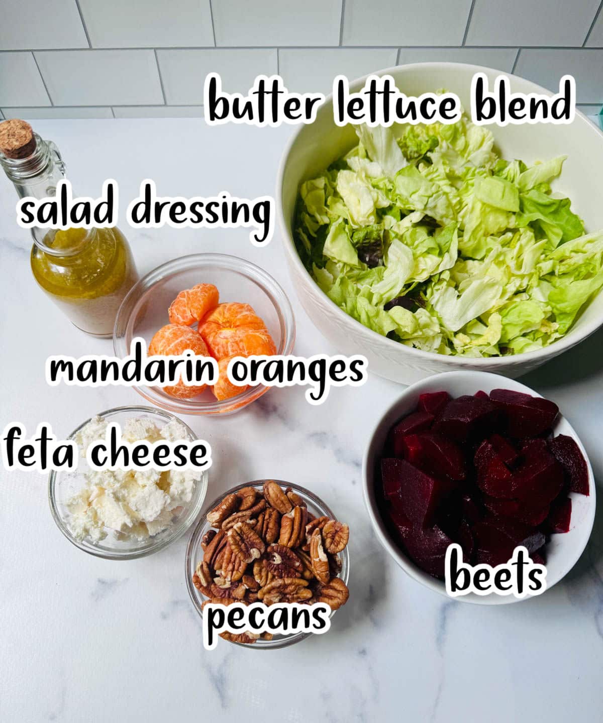 Lettuce, Mandarin orange, salad dressing bottle, beets, feta cheese, and pecans on a white counter top.