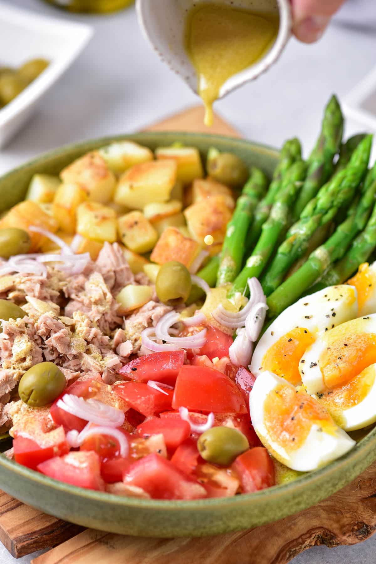 Salad with vegetables, egg, tuna, potatoes, tomatoes, and asparagus in a green bowl on a wood trivet.