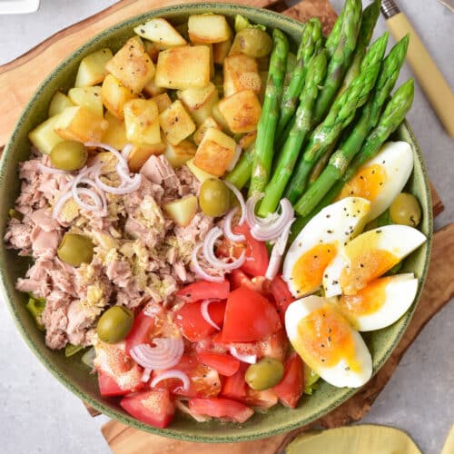Salad with vegetables, egg, tuna, potatoes, tomatoes, and asparagus in a green bowl on a wood trivet.