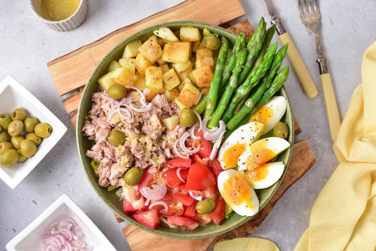 A bowl of salad containing tuna, boiled eggs, asparagus, tomato, olives, diced potatoes, and sliced onions, served on a wooden board. Forks, a napkin, and small bowls of olives and onion slices are nearby.