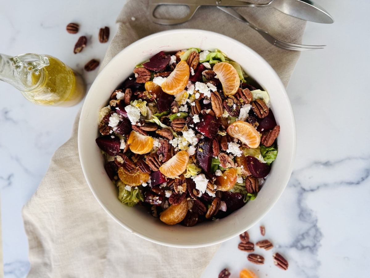 Beets, lettuce, orange sections, feta, and pecans in a white bowl on a white marble counter with cruet of salad dressing on side.
