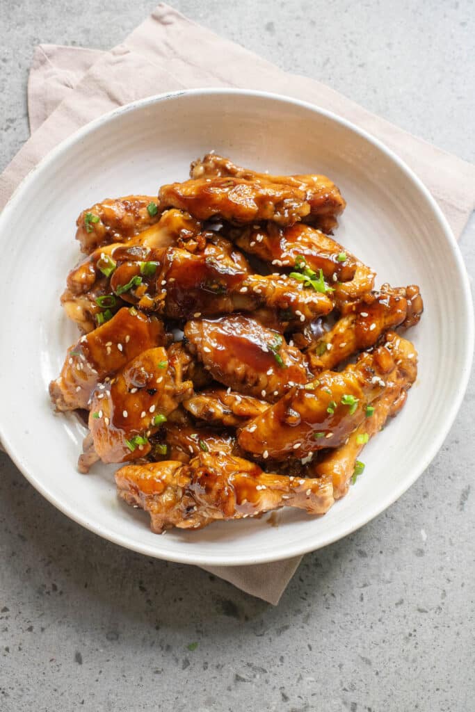 Saucy chicken wings in a white bowl with sesame seeds and green onions.