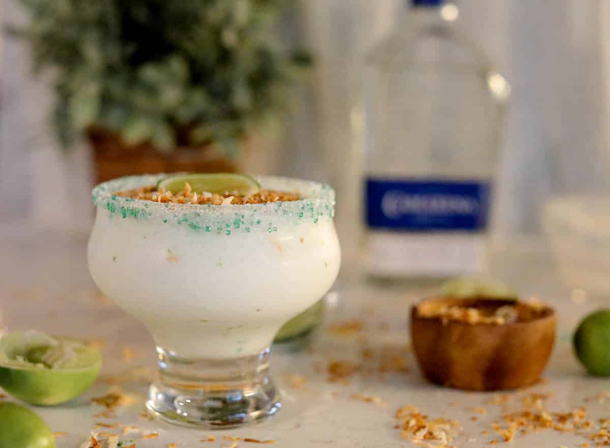 White frozen liquid in a glass with green sugar around the rim with limes and toasted coconut on the beige counter.