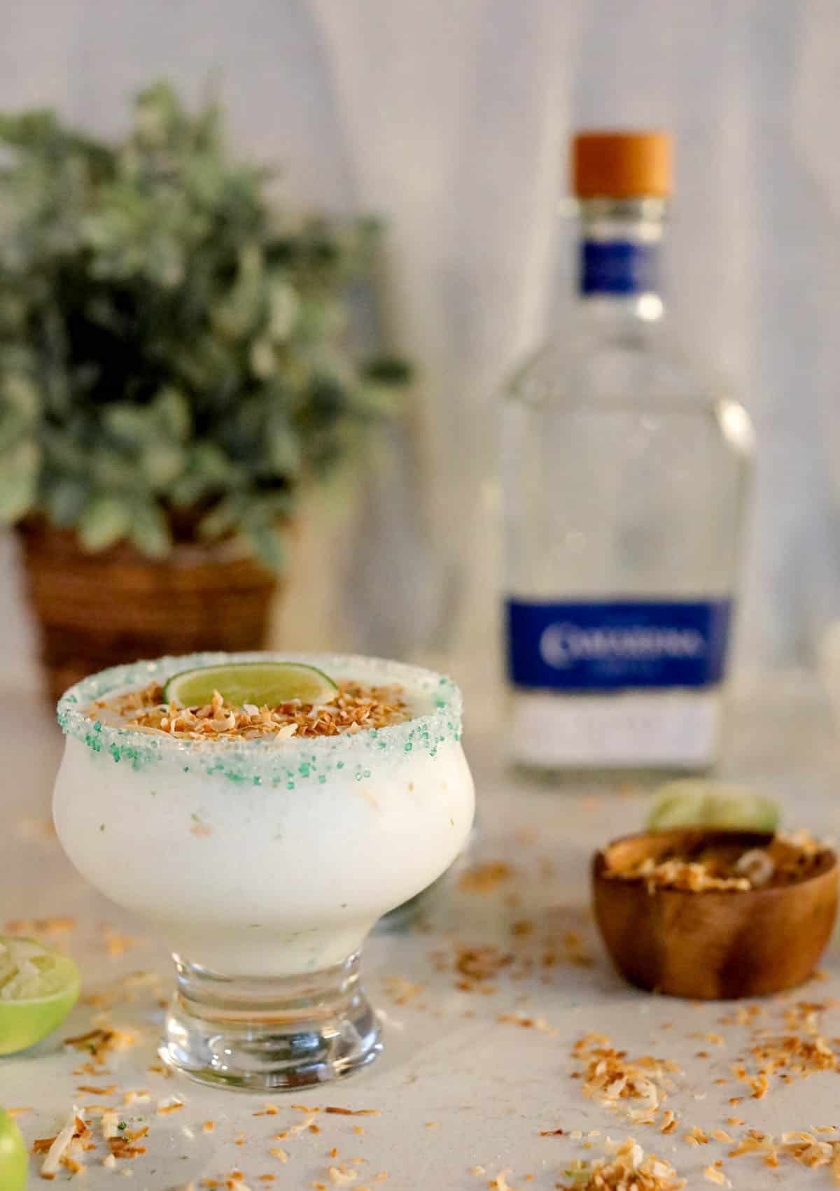 White frozen liquid in a glass with green sugar around the rim with limes and toasted coconut on the beige counter.