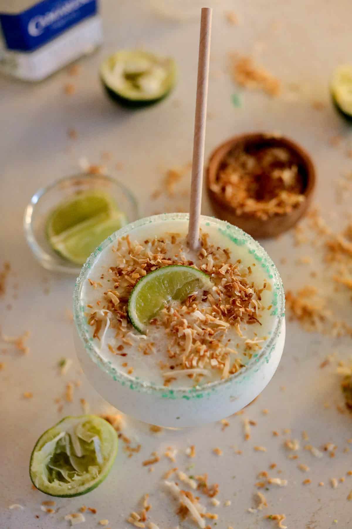White frozen liquid in a glass with a straw and green sugar around the rim with limes and toasted coconut on the beige counter.