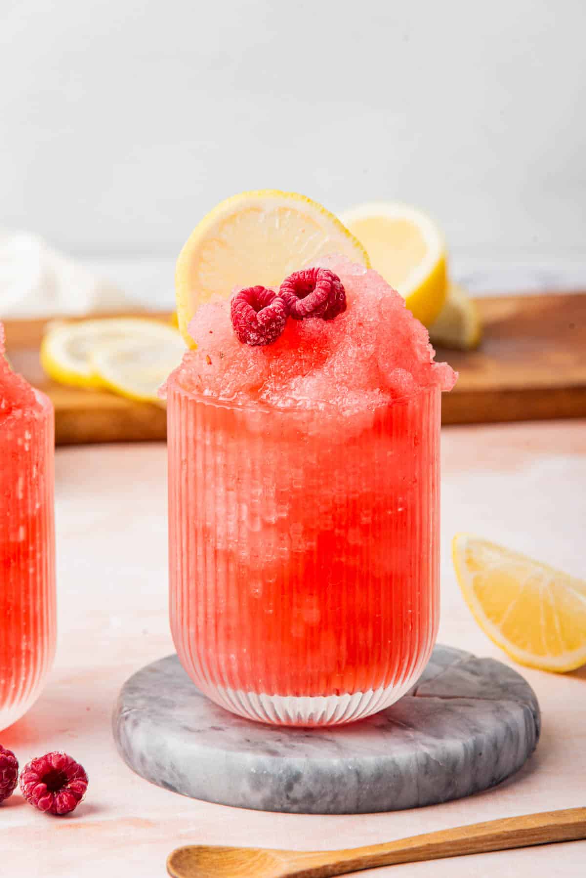 Frozen red drink in a glass with lemon and raspberries on a white counter.