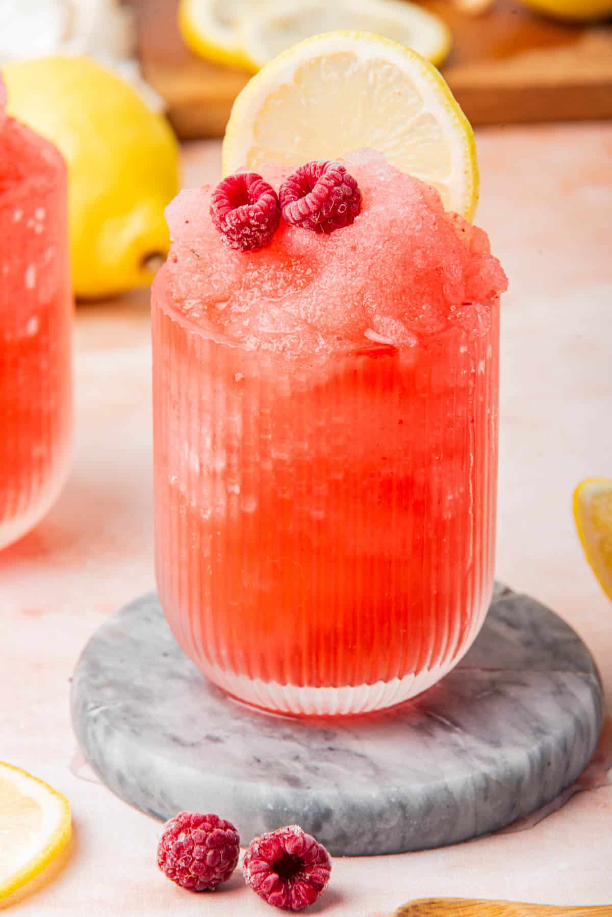 Frozen raspberry gin and tonic in a glass with lemon and raspberries on a white counter.