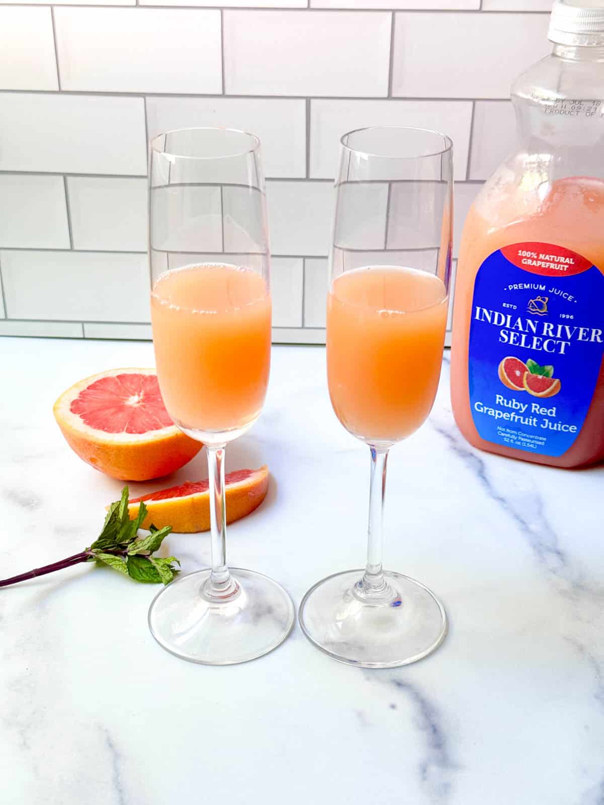 Grapefruit juice in a flute glass with fresh grapefruit, mint, and bottle of juice on white counter.