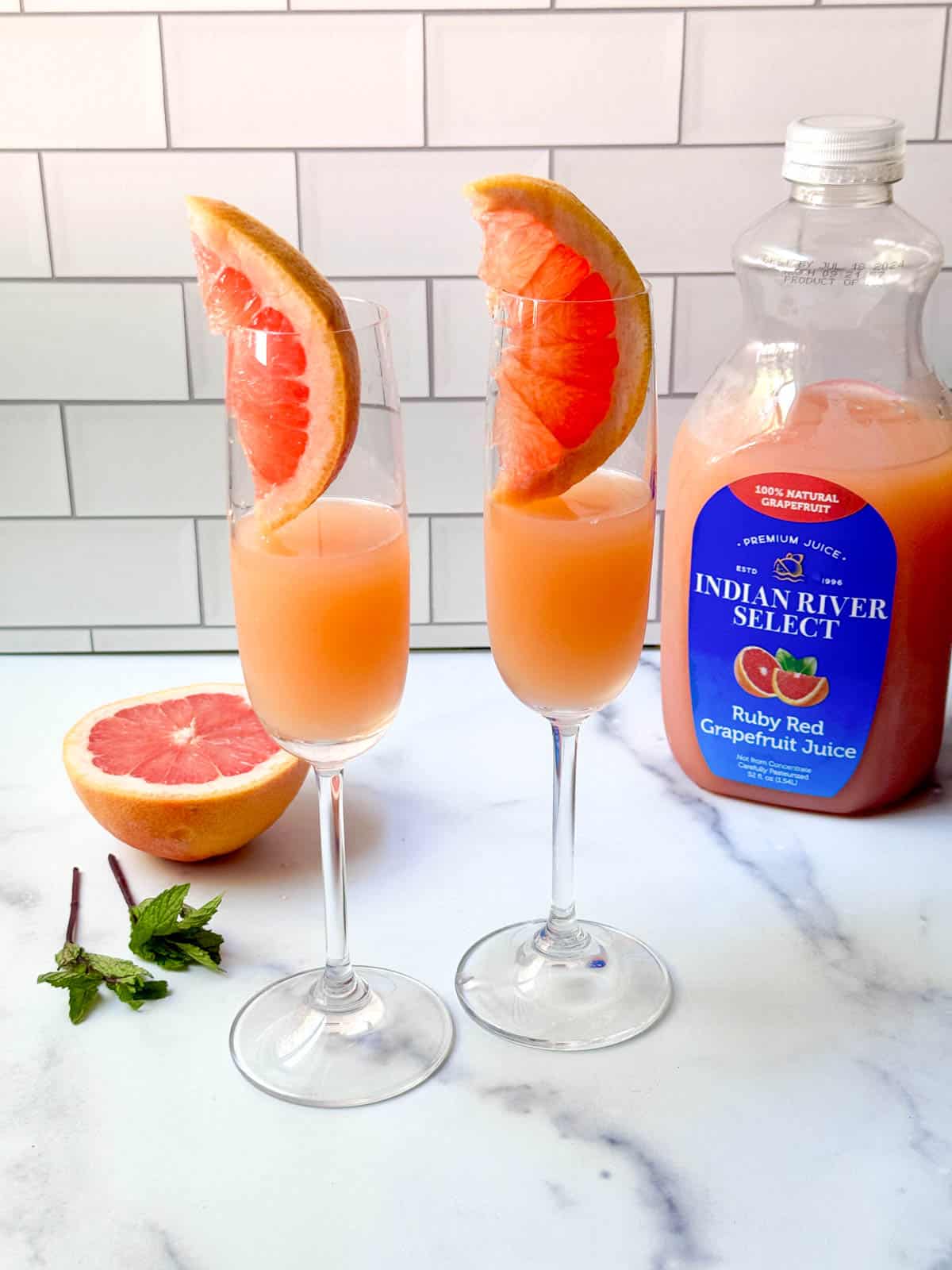 Grapefruit juice and slice of grapefruit in a flute glass on counter with fresh grapefruit, mint, and bottle of juice.
