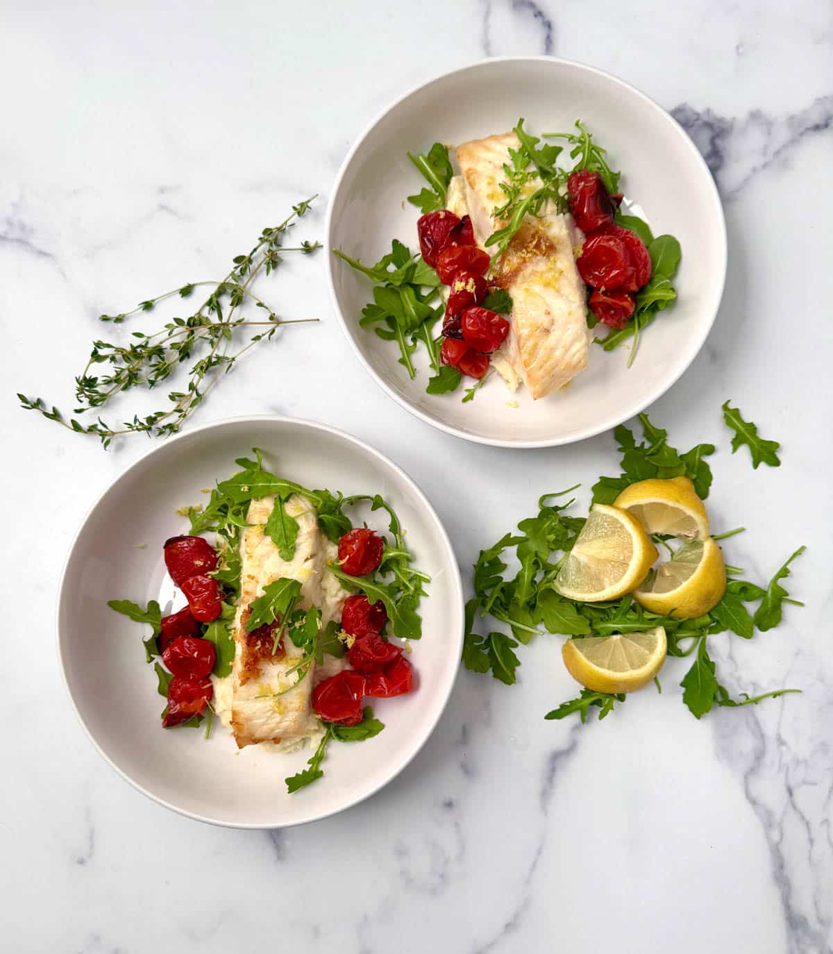 Pan seared fish in white bowls over mashed parsnips with roasted cherry tomatoes and arugula with arugula and lemon slices to the side.