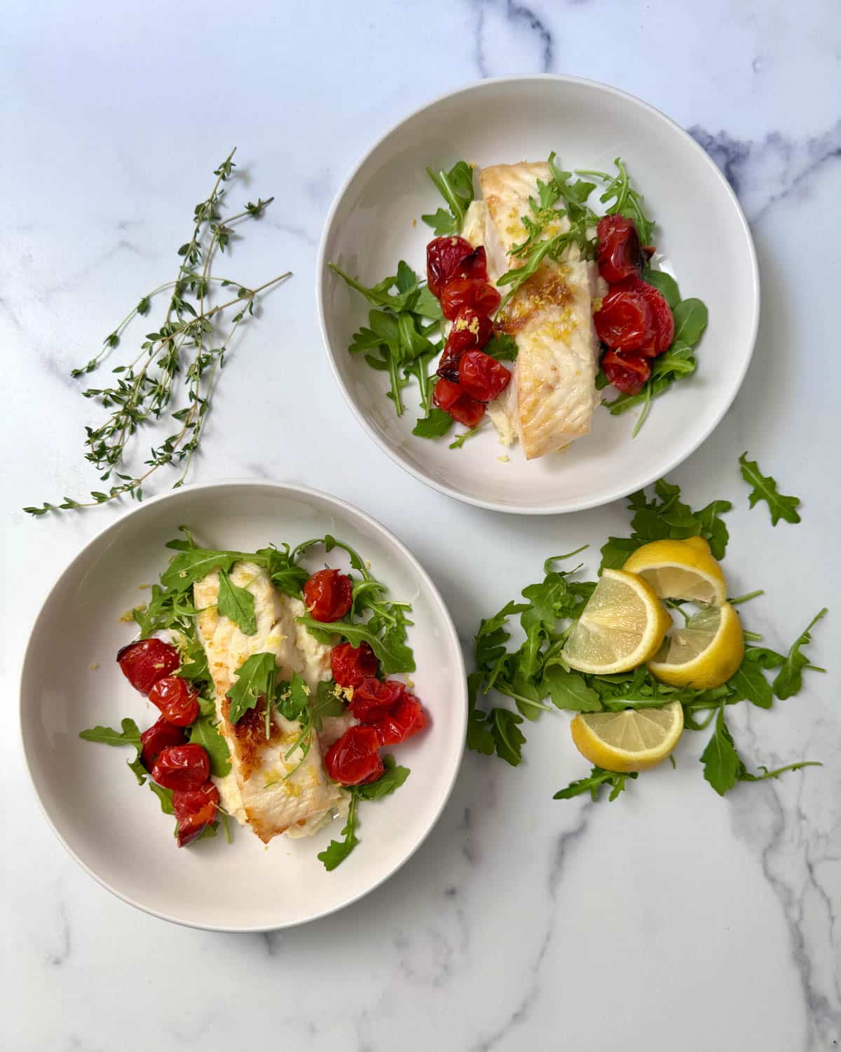Pan seared fish in white bowls over mashed parsnips with roasted cherry tomatoes and arugula with arugula and lemon slices to the side.