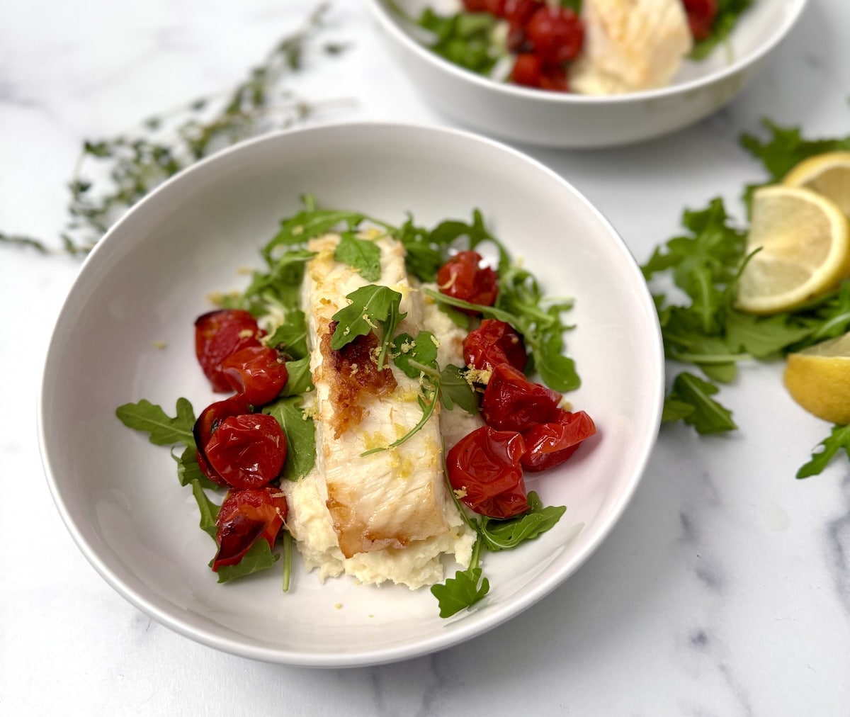 A bowl of pan-seared fish fillet on a bed of mashed parsnips, garnished with roasted cherry tomatoes, arugula, and lemon zest. Lemon wedges and extra greens are in the background.