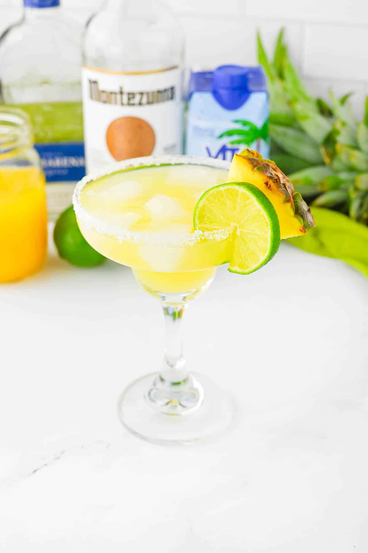 Yellow colored beverage in a margarita glass with pineapple and lime wheel on salted rim and other ingredients in background.