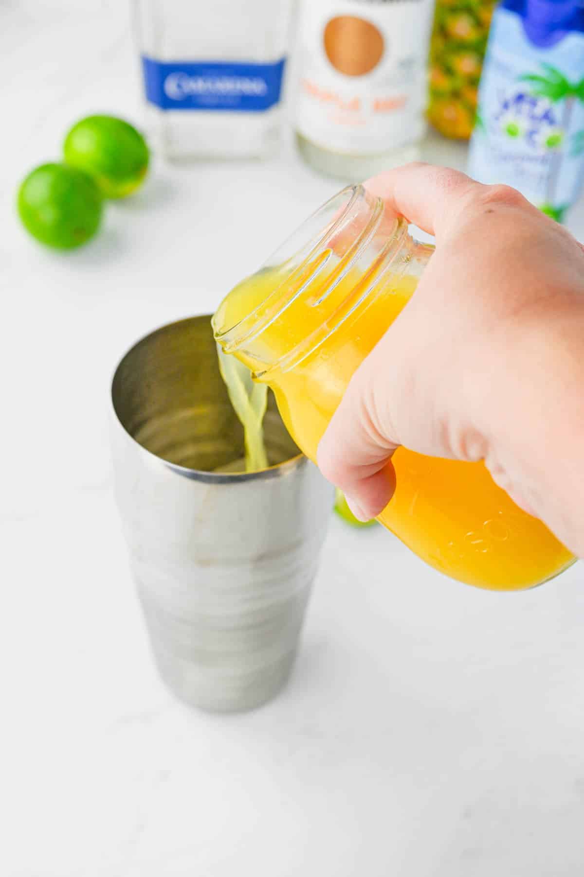 Pouring pineapple juice into a cocktail shaker with bottle of tequila, triple sec, coconut water, and fresh pineapple in background.