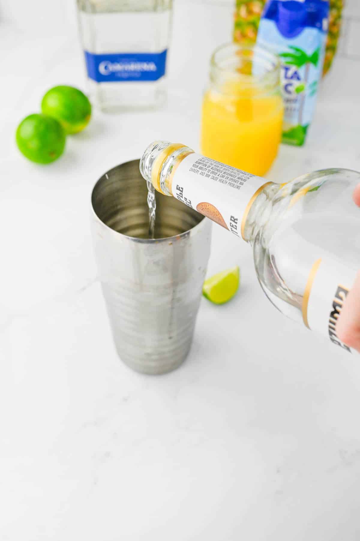 Pouring triple sec into a cocktail shaker with other ingredients in background on counter.