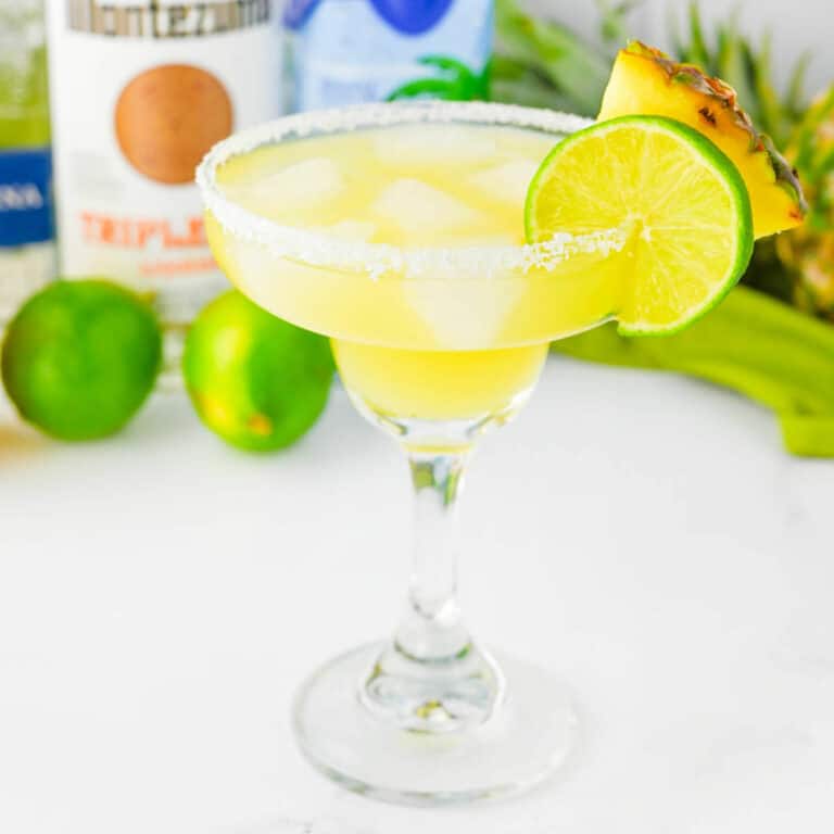 Yellow colored beverage in a margarita glass with pineapple and lime wheel on salted rim and other ingredients in background.