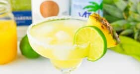 Yellow colored beverage in a margarita glass with pineapple and lime wheel on salted rim and other ingredients in background.