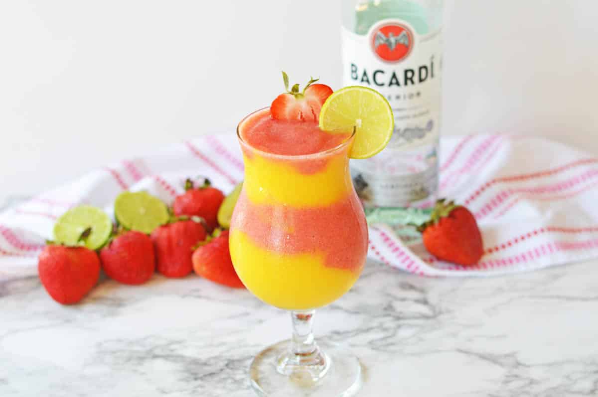 Frozen strawberry and mango daiquiri in a hurricane glass with a bottle of rum in background.
