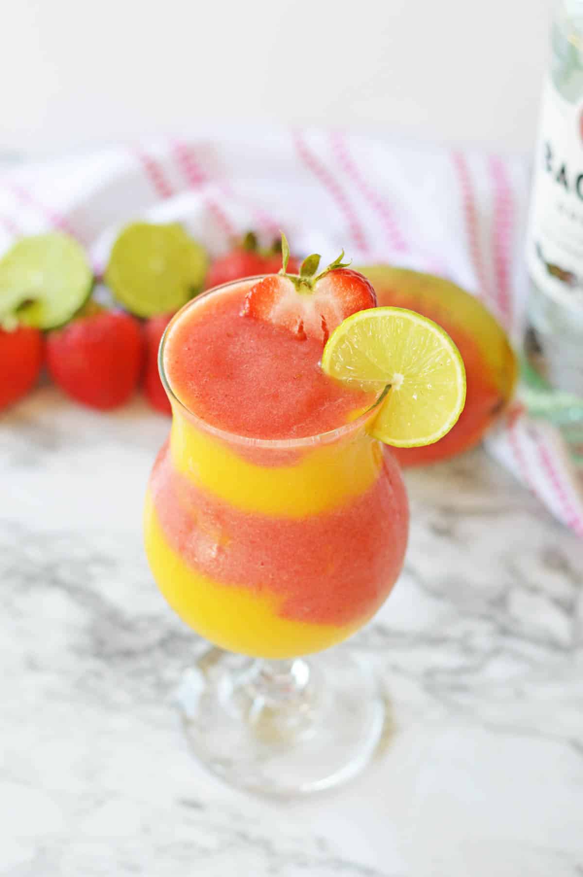 Strawberry mango daiquiri in a hurricane glass with a strawberry and lime slice and fruit in background.