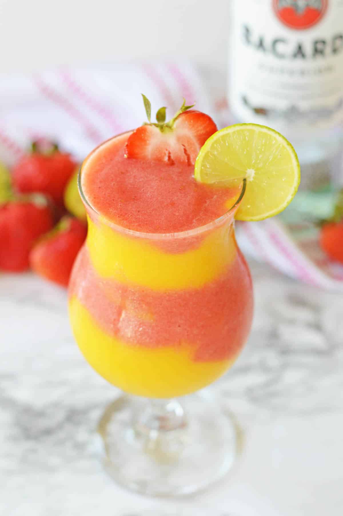 Frozen strawberry and mango layers in a hurricane glass with a strawberry and lime wheel garnish.