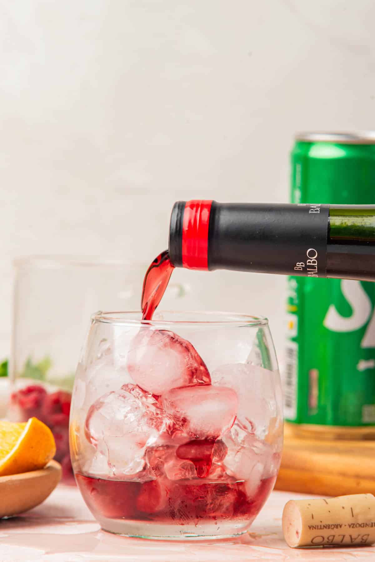 Pouring red wine over ice in a cocktail glass with fruit and can of Sprite in background.