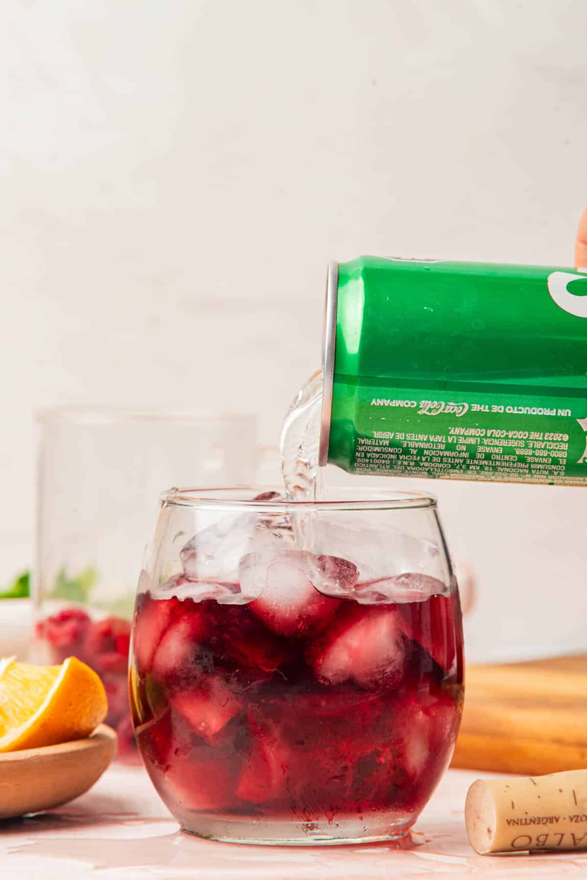 Adding Sprite to red wine over ice in a cocktail glass with fruit and can of Sprite in background.