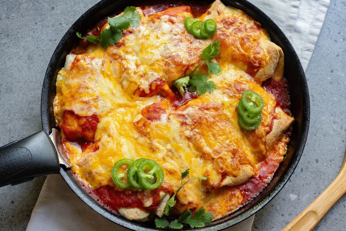 A burrito topped with enchilada sauce and cheese, jalapenos, and cilantro in a cast iron pan.
