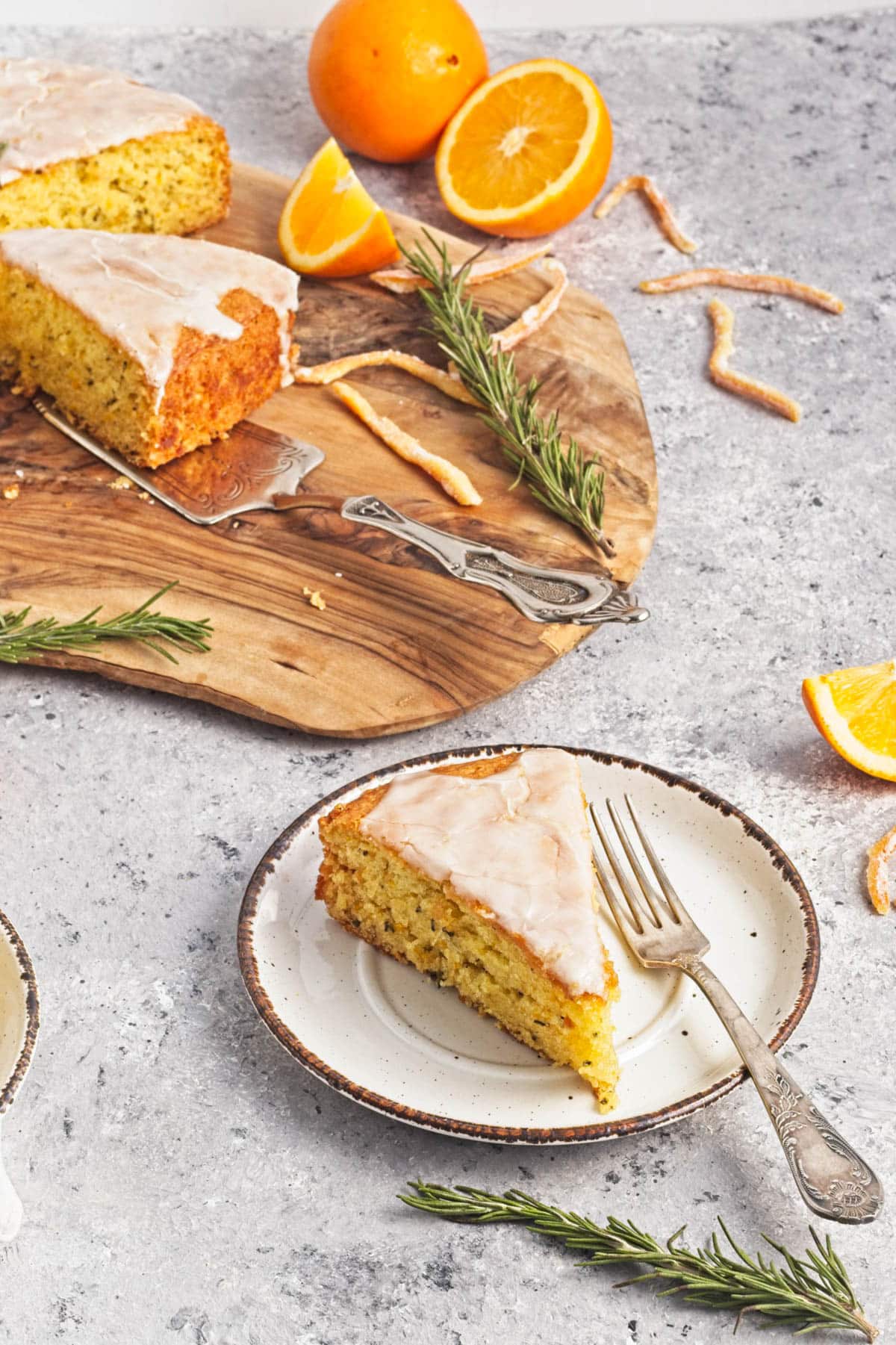 Orange olive oil cake with rosemary topped with icing and orange zest with oranges to the side.