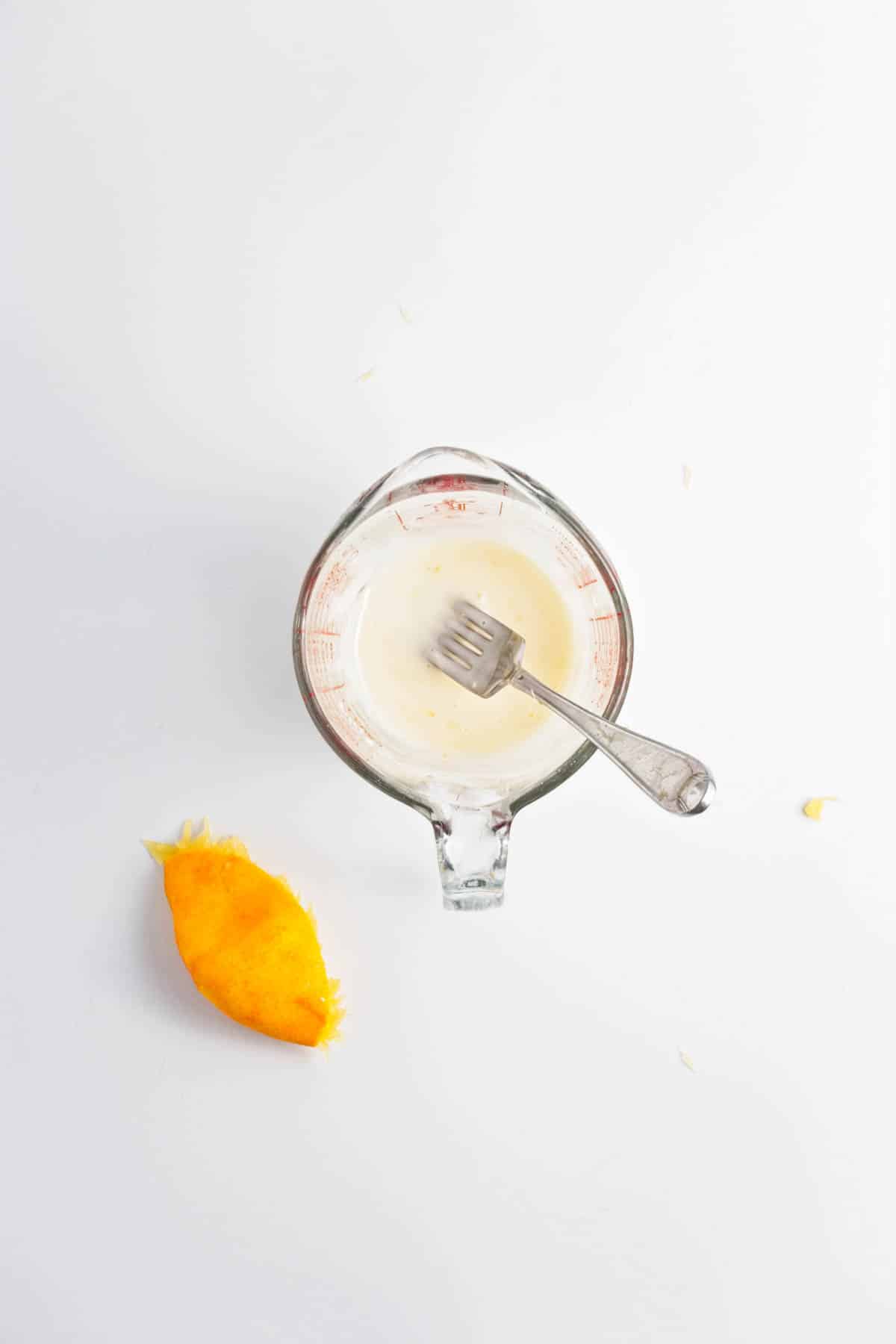 Orange juice and powdered sugar icing in a measuring cup with a fork and orange peel to the side.