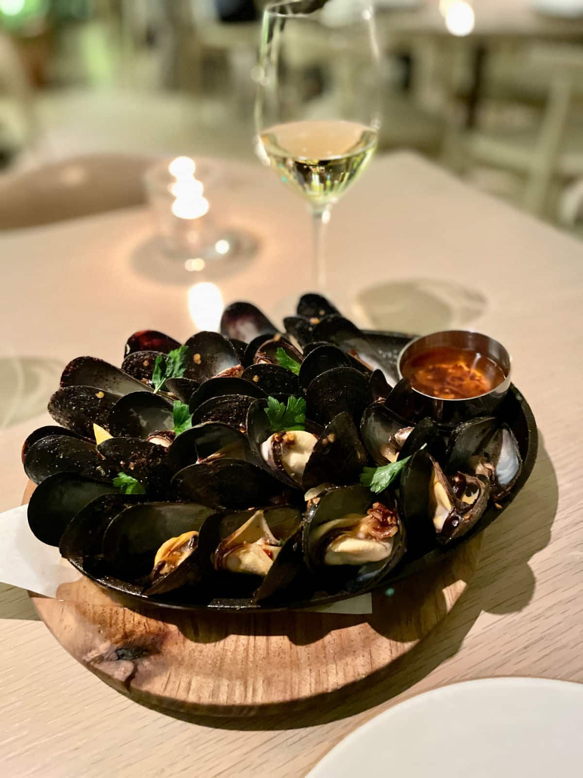 Mussels in a black bowl on a wood plate with chili oil and cilantro with a glass of wine in background.