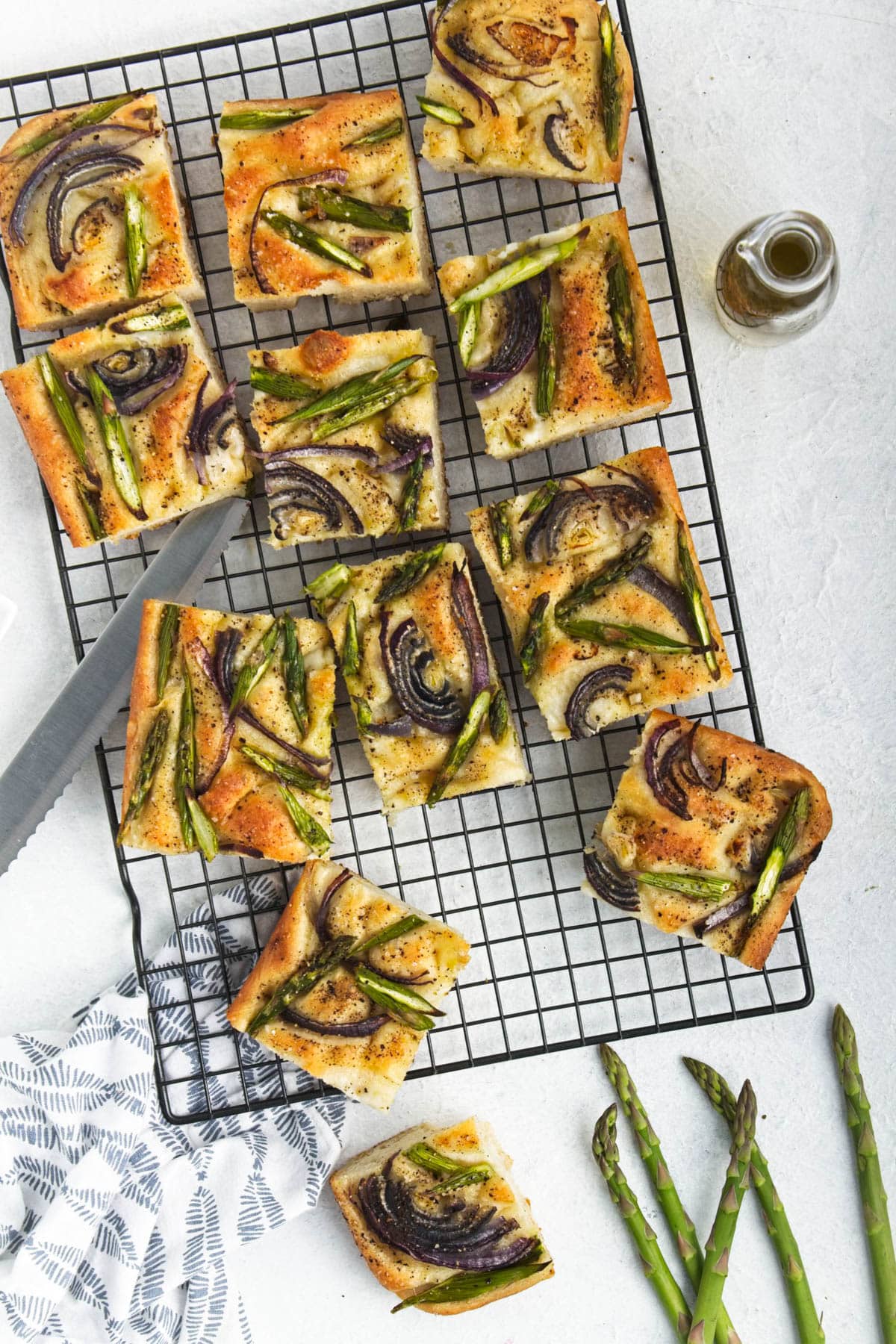 Asparagus and onion focaccia on a cooling rack cut into squares with bottle of oil on side.