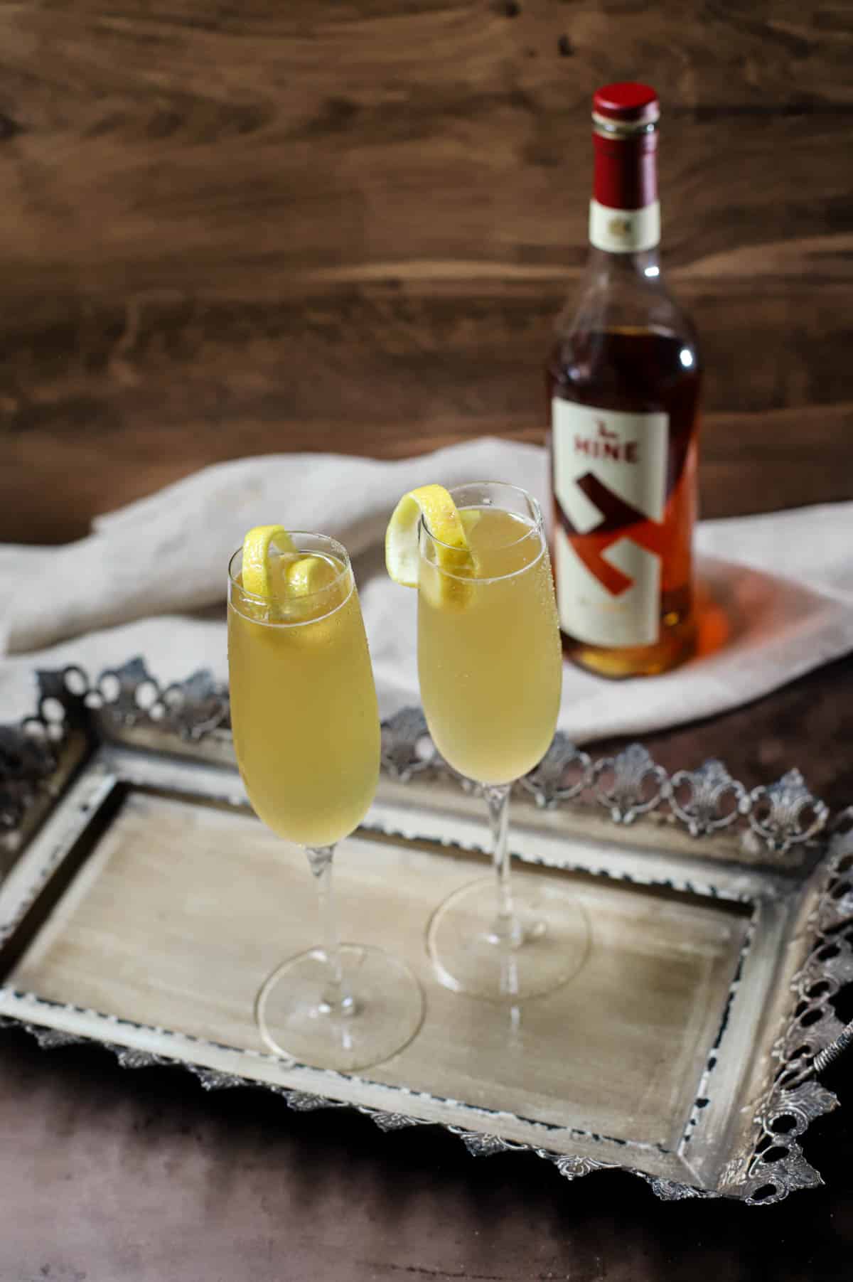 French 75 cocktail on a tray with bottle of cognac in back on dish towel.