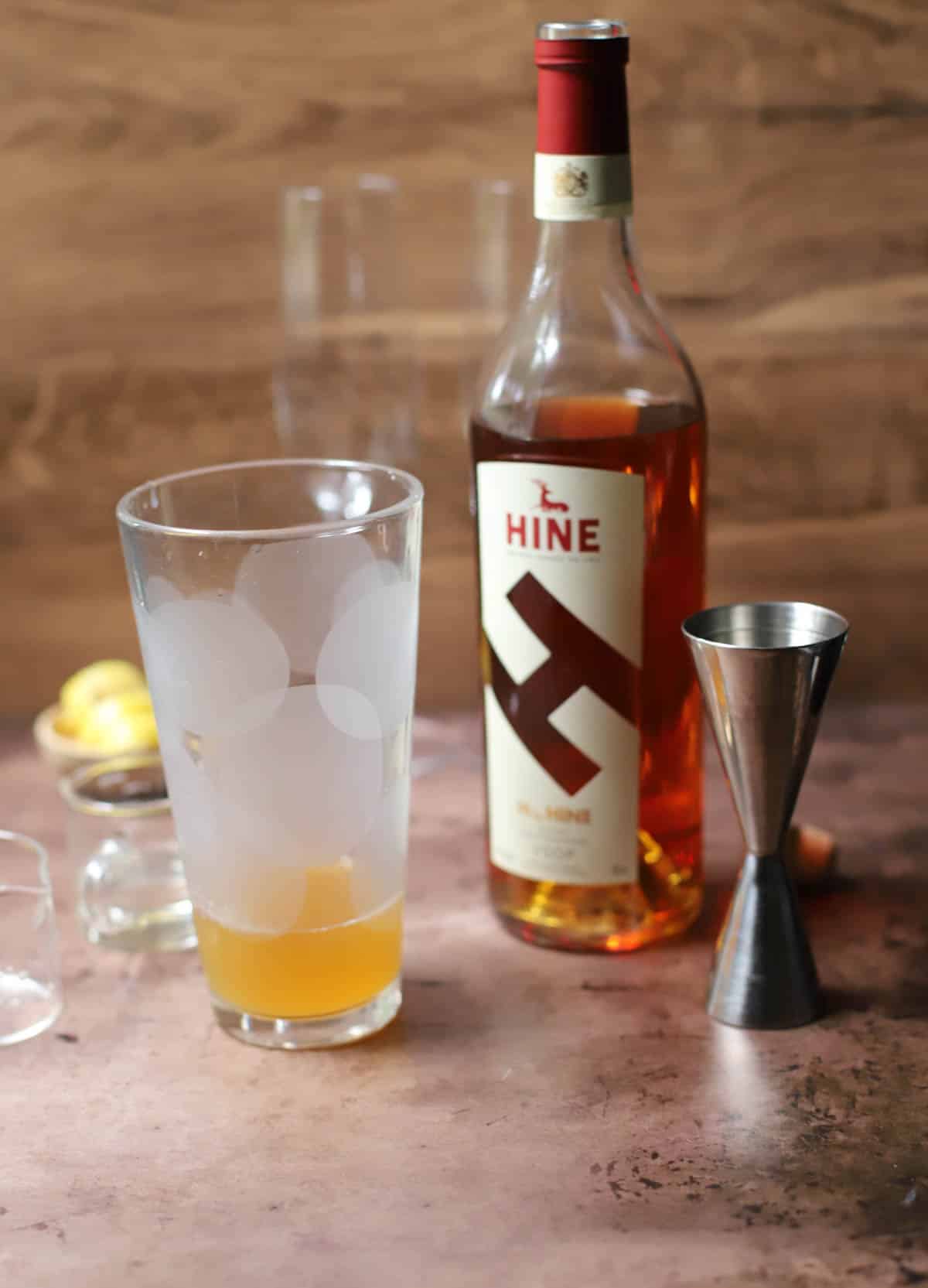 Cocktail shaker glass with cognac, lemon juice, and simple syrup with bottle and stainless jigger to the side.
