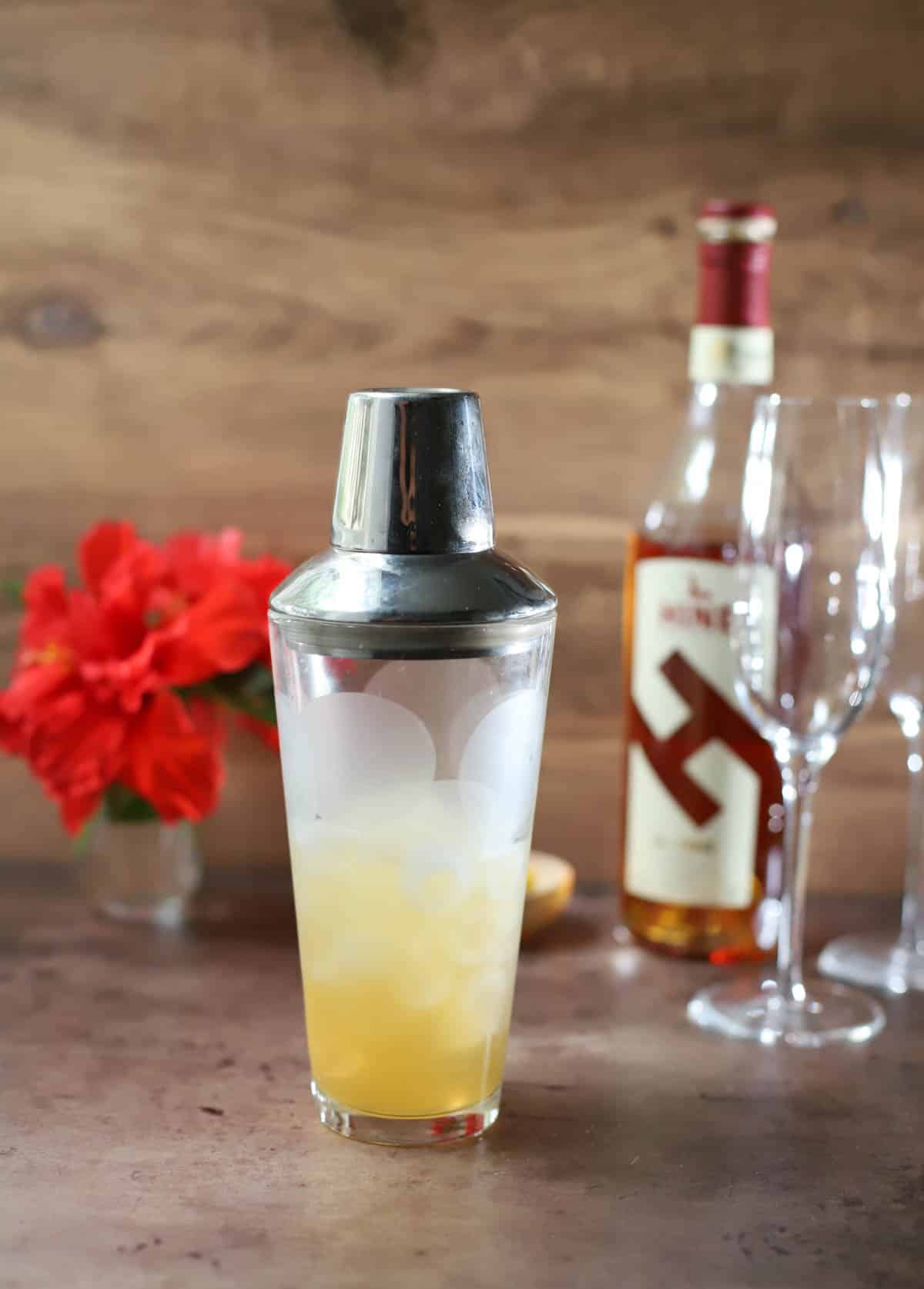 Shaker of French 75 cocktail on dark table with wood background with bottle and flowers behind.