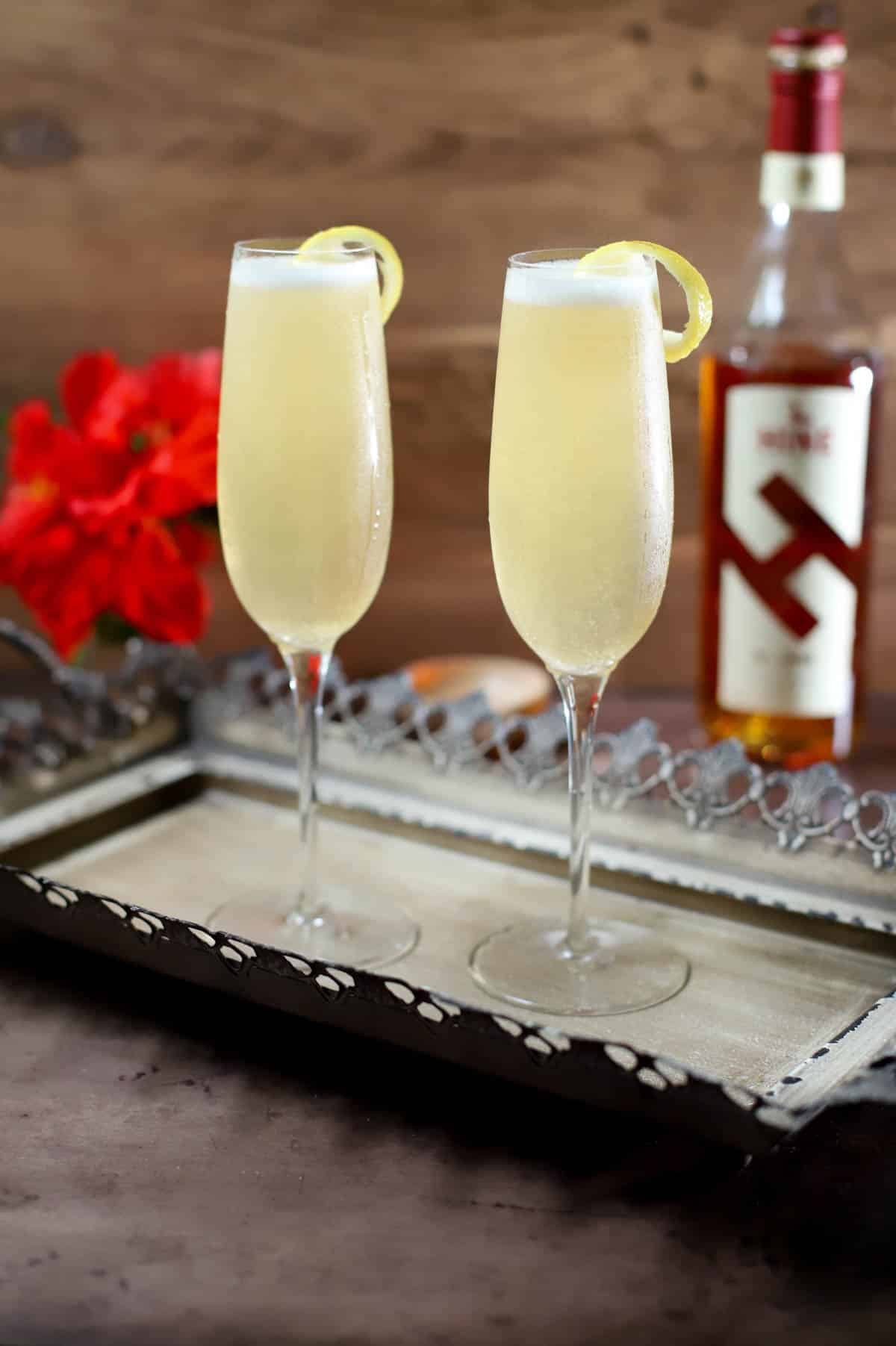 French 75 cocktail on a tray with flowers and bottle of cognac in back.