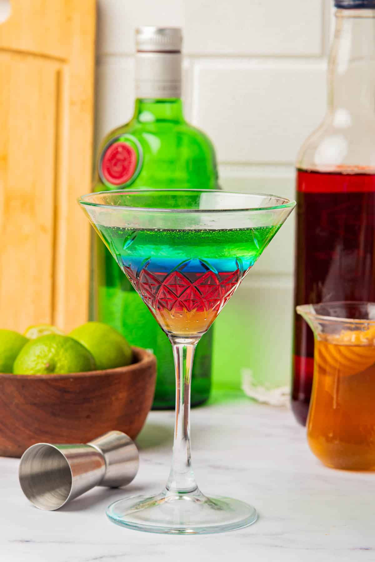 Martini glass with liquid in the colors of the Olympic Rings with a jigger on side, limes, bottles, and honey in background.