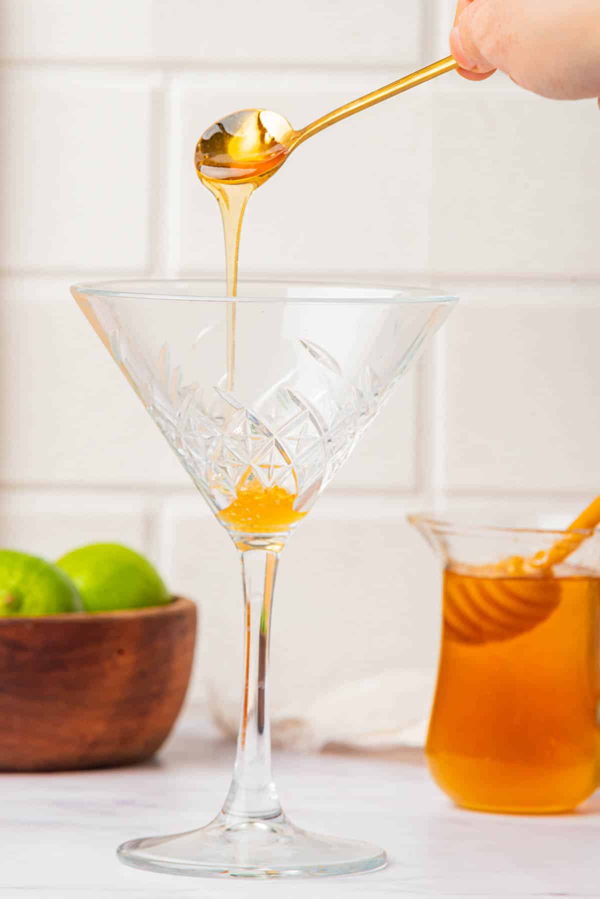 Pouring honey into a martini glass with honey and limes in background.