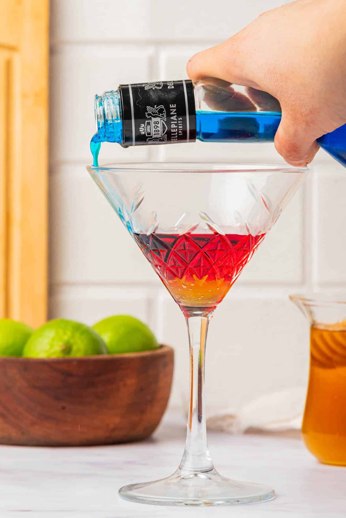 Pouring blue liquid into a martini glass over orange and red liquid with bowl of limes and cup of honey in background.