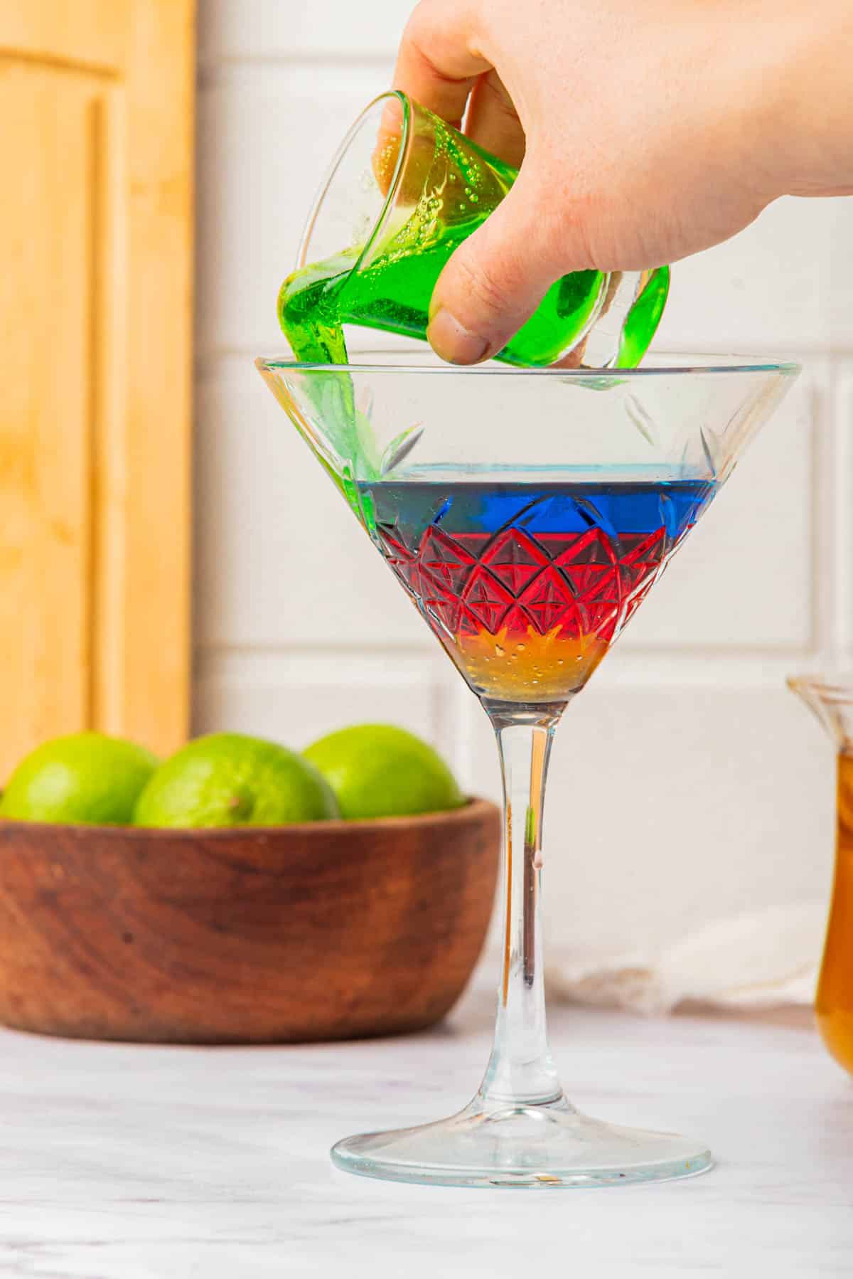 Pouring green liquid into a artini glass with liquid in the colors of the Olympic Rings with limes and a cup of honey in background.