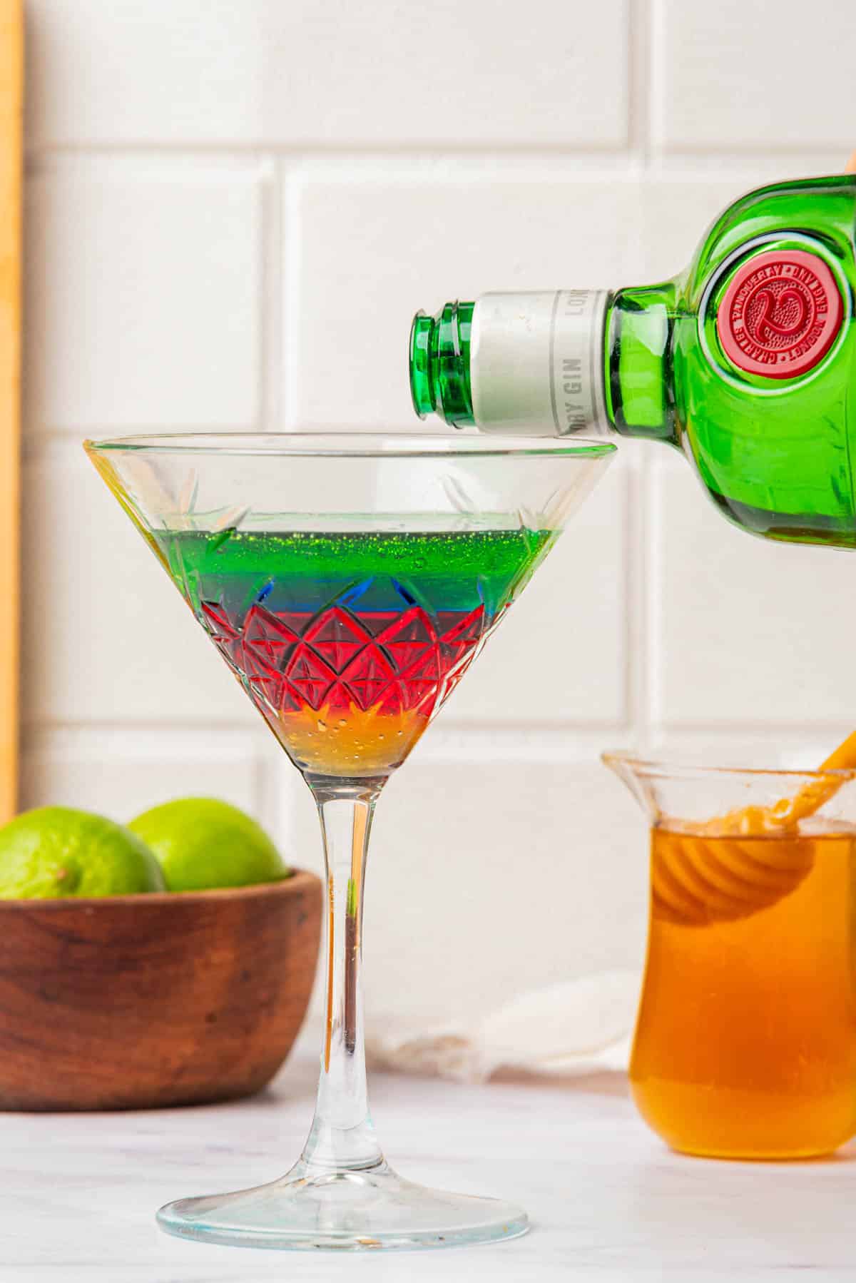 Pouring gin into a martini glass with liquid in the colors of the Olympic Rings with a jigger on side, limes, bottles, and honey in background.