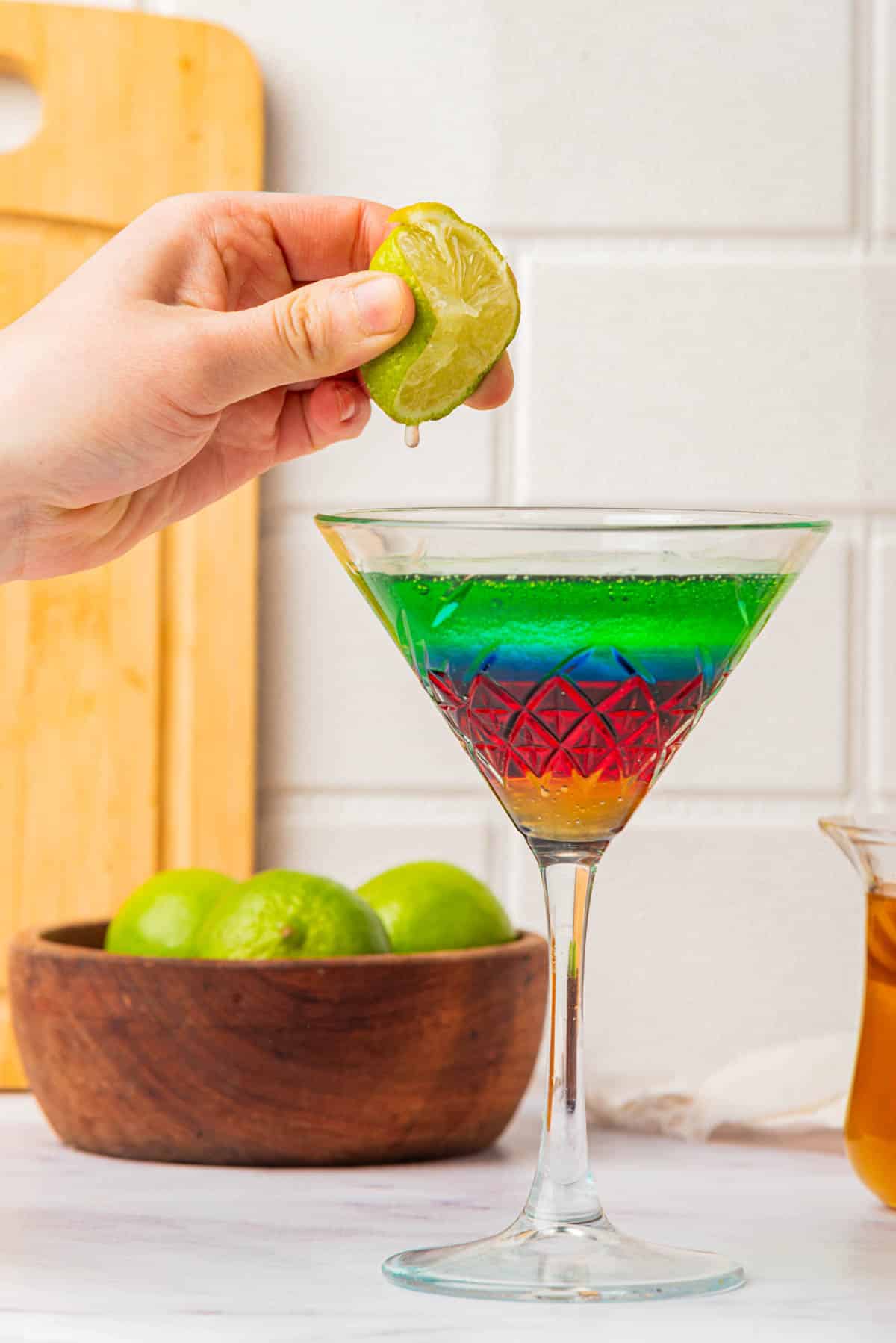 Squeezing lime into a martini glass with liquid in the colors of the Olympic Rings with a jigger on side, limes, bottles, and honey in background.